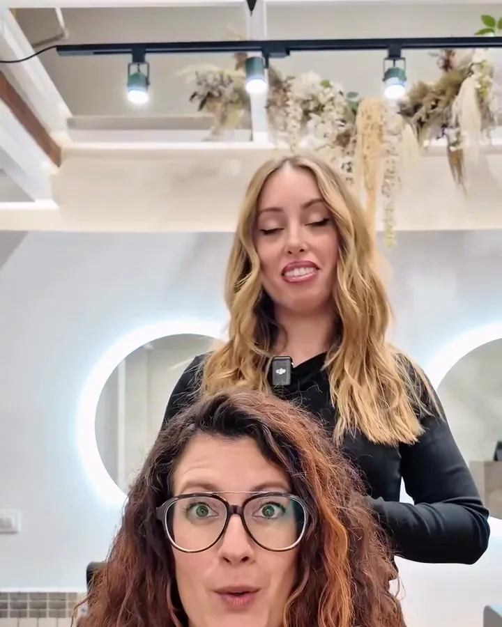 Ari sitting in front of the salon mirror before her first curly cut, with her curly hair loose and undefined, and the stylist standing behind her. Still from the salon's video.