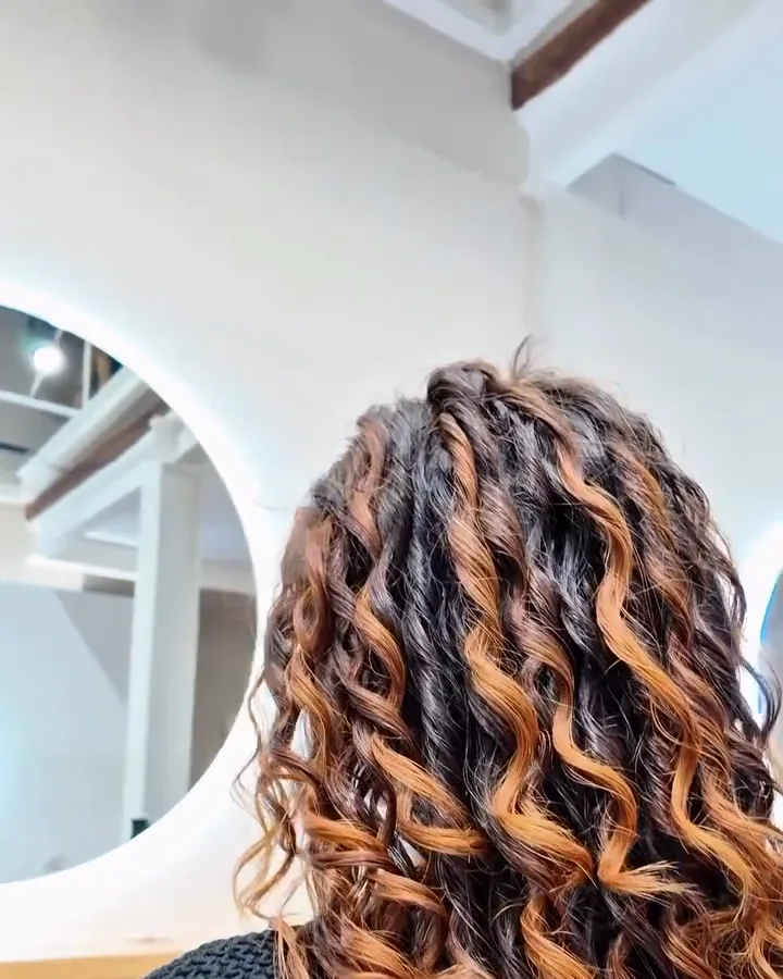 Ari's hair seen from behind with defined curls and the copper highlights visible. Still from the salon's video.