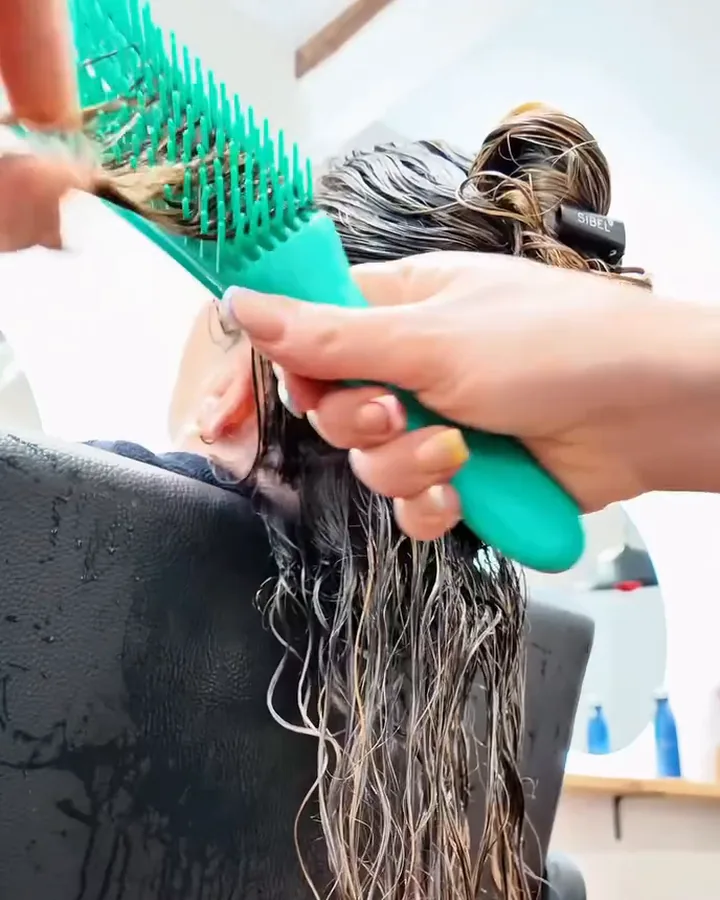 A hand combs Laura's wet hair with a teal, wide-toothed brush, seen from the side without showing her face. Still from the salon's video.